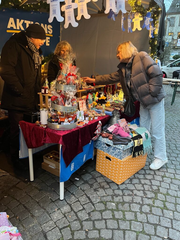 A woman in a blue jacket shops at an outdoor market stall decorated with lights and holiday items, speaking to two vendors behind a table filled with handmade crafts, gifts, and winter accessories.