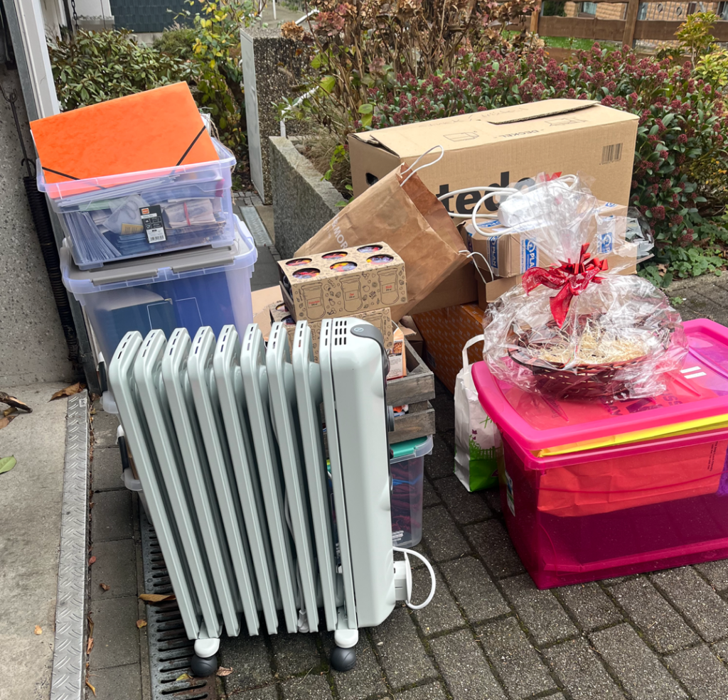 A collection of household items and supplies, including a portable radiator, plastic storage boxes, cardboard boxes, shopping bags, and a wicker gift basket, arranged on a paved outdoor area next to greenery.
