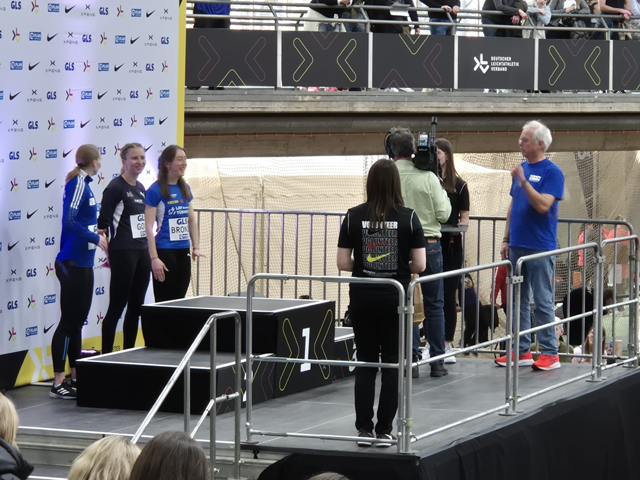 Three female athletes stand near a podium as a man in blue prepares to present awards. A camera crew films the scene, and spectators watch from behind a barrier and on the balcony above.