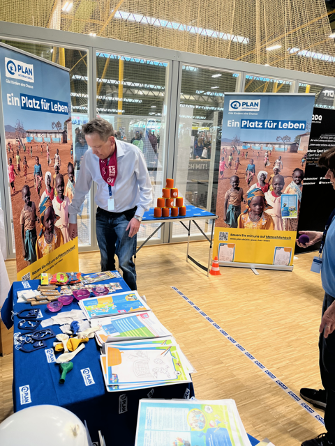 A man stands next to a table with educational materials, pamphlets, and toys at an indoor event. Behind him are posters with images of children and the German text Ein Platz für Leben.