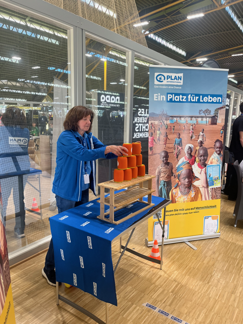 A woman stacks orange cans on a wooden frame at an indoor event booth. Behind her is a banner for Plan International with images of children and the text Ein Platz für Leben.