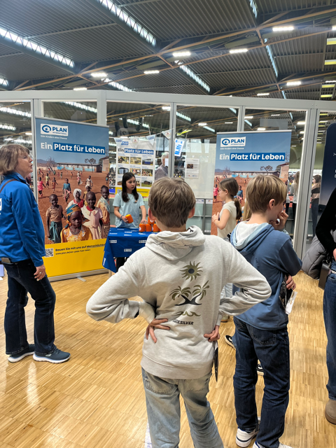 Children and adults stand in front of a booth with PLAN banners at an indoor event. The banners read Ein Platz für Leben and display images of children. The floor is wooden and the setting is a large hall.