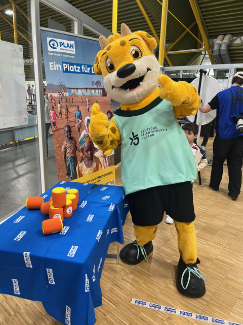 A cheerful yellow mascot in a light green shirt gives a thumbs up at an event booth, with colorful toy blocks on a blue table and a child partially visible behind. A banner and posters are displayed in the background.
