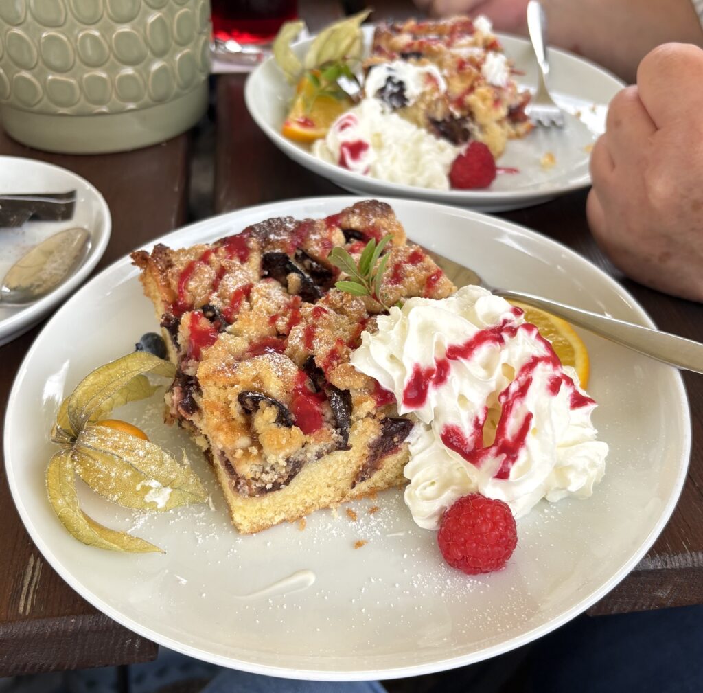 Ein Teller mit einem Stück Obst-Crumble-Kuchen, Schlagsahne, einer Himbeere und einer Physalis. Im Hintergrund ist eine Hand mit einer Gabel zu sehen, in der Nähe eines weiteren Tellers mit einem ähnlichen Dessert.
